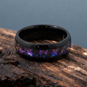 Könnte beinhalten: Ein schwarzer Ring mit einem galaktischen Inlay in Lila-, Blau- und Goldtönen. Der Ring ist auf einer dunklen Holzoberfläche platziert, die seine polierte Oberfläche und sein einzigartiges Design hervorhebt.