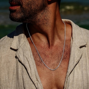 Puede incluir: Un collar de cadena de plata usado por una persona con una camisa de lino marrón claro.