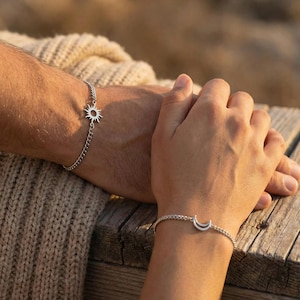 May include: Two silver bracelets, one with a sun charm and the other with a crescent moon charm, are displayed. The bracelets are on a chain link style. The background is blurred, with a wooden surface and a beige sweater.