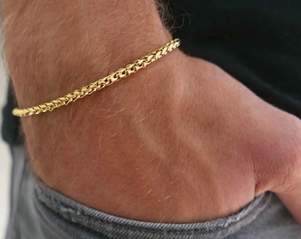 May include: A gold-colored chain bracelet with a woven design. The bracelet is worn on a wrist, with the person wearing a black shirt and gray jeans. The bracelet is a simple, elegant accessory.