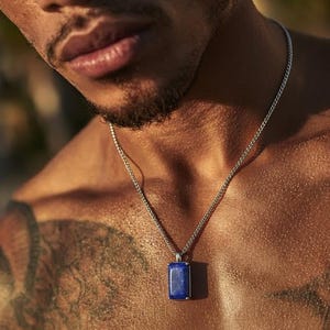 Pode incluir: Um colar de corrente prateada com um pendente retangular azul escuro. O pendente é emoldurado em prata. O colar é usado sobre um peito nu, com tatuagens visíveis no ombro.