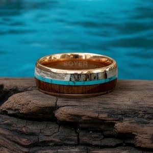 May include: A gold-toned ring with the word "SOYER" engraved on the inner band. The ring features a wood grain band, a turquoise band, and a white band. The ring is displayed on a piece of wood.