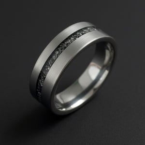 Puede incluir: Un anillo plateado con una incrustación oscura y texturizada. El anillo tiene un acabado de metal cepillado y un interior pulido. La incrustación parece estar compuesta por pequeños fragmentos oscuros. El anillo está sobre un fondo oscuro.