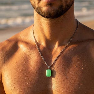 May include: A silver chain necklace with a rectangular, light green pendant. The pendant is approximately 0.75 inches in length and 0.5 inches in width. The necklace is worn on a person's bare chest, with a beach and ocean in the background.