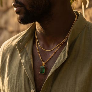 May include: A gold-colored necklace with a rectangular emerald-green pendant, layered with a thinner gold chain. The jewelry is worn over a light green button-down shirt.