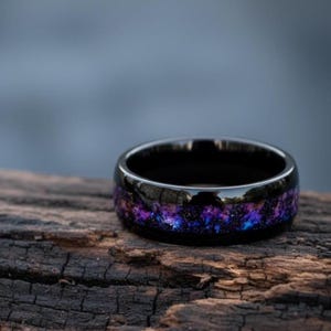May include: A black ring with a galaxy design. The ring features a polished black band with a vibrant inlay of purple, blue, and white, resembling a starry night sky. The ring is set on a dark wooden surface.