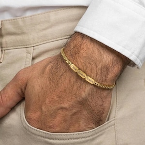 Peut inclure: Un bracelet doré avec un motif de chaîne tressée. Le bracelet comporte trois plaques nominatives rectangulaires gravées des noms "Chloe", "Annabelle" et "Scarlett". Le bracelet est porté au poignet, avec une chemise blanche et un pantalon beige en arrière-plan.