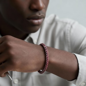 May include: A burgundy and silver chain bracelet is shown on a person's wrist. The bracelet features a series of interconnected links, with a burgundy material woven through the silver links. The person is wearing a light-coloured shirt.