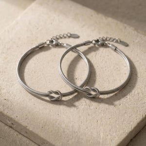 May include: Two silver-colored bracelets with a knot design. The bracelets are made of a thin, flexible material and have adjustable clasps. The bracelets are displayed on a light beige surface.