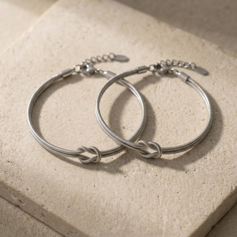 May include: Two silver-colored bracelets with a knot design. The bracelets are made of a thin, flexible material and have adjustable clasps. The bracelets are displayed on a light beige surface.