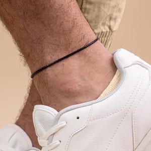 以下が含まれることがあります： 黒いアンクレットが足首に着用され、白いスニーカーが前景にあります。アンクレットは薄く、質感のある素材でできているようです。スニーカーは白で、白いソールと靴紐が付いています。