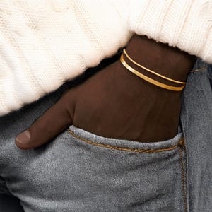 May include: A gold bracelet with a simple, flat band design. The bracelet is worn on the wrist. The person is wearing a white sweater and blue jeans. The bracelet is a stylish accessory.