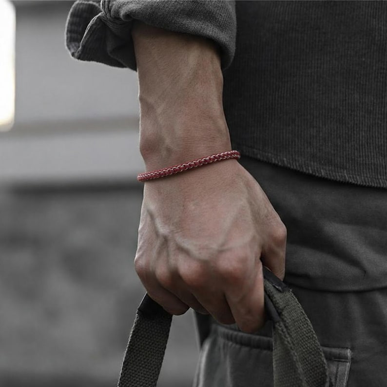 May include: A braided red bracelet is worn on a wrist. The bracelet is made of a woven material and appears to be a single strand. The person is wearing a grey long-sleeve shirt and holding a green bag.