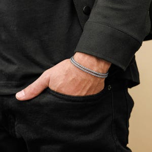May include: A silver-coloured braided bracelet is worn on a wrist. The bracelet is made of two strands of braided metal. The person is wearing a black jacket and black trousers. The background is a neutral beige colour.