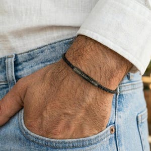 Op de afbeelding: Zwart gevlochten armband met een rechthoekige metalen plaat gegraveerd met de namen "Emma | Adam | Lanna". De armband wordt om de pols gedragen, de persoon draagt een wit overhemd en een blauwe jeans.