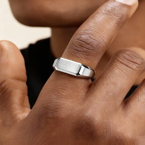 Op de afbeelding: Een zilverkleurige ring met een rechthoekige, geborstelde metalen bovenkant en geometrische zijkanten. De ring wordt aan een vinger gedragen en toont zijn gepolijste afwerking en moderne design. De ring is een sieraad.