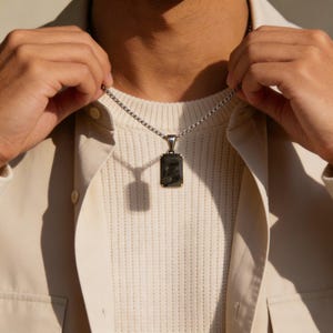 Puede incluir: Un collar de cadena plateada con un colgante rectangular. El colgante tiene una piedra oscura y texturizada engastada en un marco dorado. El collar está siendo sostenido por una persona.