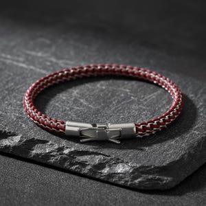 May include: A burgundy and silver braided bracelet with a stainless steel clasp. The bracelet is displayed on a dark grey, textured surface. The jewellery is a stylish accessory.