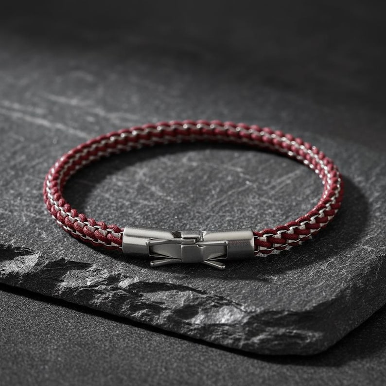 May include: A burgundy and silver braided bracelet with a stainless steel clasp. The bracelet is displayed on a dark grey, textured surface. The jewellery is a stylish accessory.