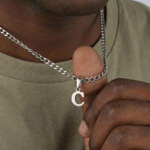 Puede incluir: Collar de cadena plateada con un colgante en forma de la letra "C". El colgante está suspendido de una pequeña fianza. La cadena es de estilo curb. El collar lo sostiene una persona.