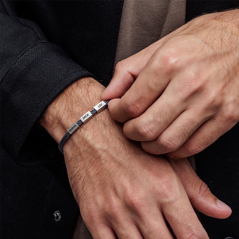 May include: A black and gray braided bracelet with silver metal beads. The beads are engraved with the names "Emma", "Jack", and "Liv".