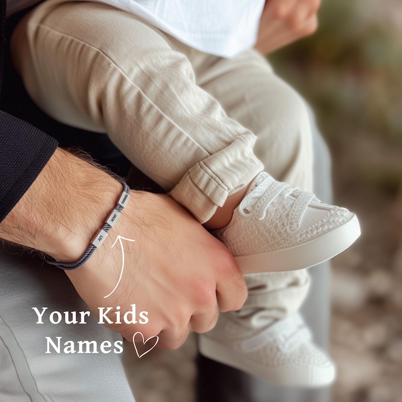 May include: A man wearing a bracelet with the names of his children. The bracelet is made of gray cord with silver beads. The man is holding a child in his arms. The child is wearing white shoes.