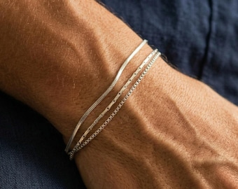 Pode incluir: Um conjunto de três pulseiras de tom prateado no pulso. O conjunto inclui uma corrente de cobra lisa, uma corrente de elos retangulares e uma corrente de caixa. Cada pulseira apresenta um acabamento metálico e um design fino e delicado.