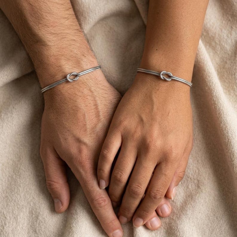 May include: Two silver bracelets, each featuring a knot design at the center. The bracelets are worn on wrists, with the hands clasped together. The bracelets are made of a thin, metallic material.