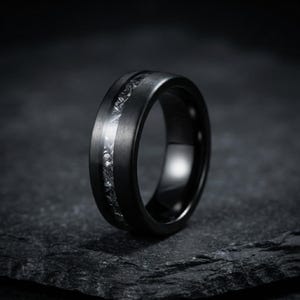 Peut inclure: Une bague noire mate dotée d'une incrustation centrale au fini métallique argenté texturé. L'anneau présente une texture de surface brossée et lisse ainsi qu'un profil arrondi, mettant en valeur un contraste net entre l'extérieur sombre et l'incrustation claire.