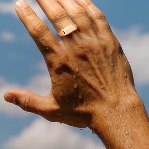Puede incluir: Un anillo de sello dorado con una cara rectangular se lleva en una mano. La mano está mojada con gotas de agua, y el fondo es un cielo azul con nubes blancas. El anillo es el punto focal de la imagen.