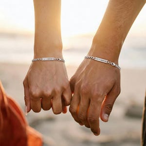 Puede incluir: Dos pulseras de cadena plateadas con placas rectangulares con nombres, cada una grabada con números romanos. Las pulseras están en muñecas, con las manos entrelazadas. El fondo es una escena de playa borrosa con una luz cálida y dorada.