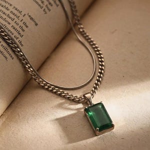 May include: A silver-toned necklace with two chains. The lower chain is a curb chain, and the upper chain is a snake chain. A rectangular, emerald-green pendant hangs from the curb chain. The necklace rests on a book.