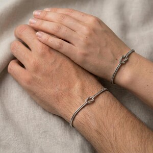May include: Two silver bracelets, each featuring a knot design, are worn on wrists. The bracelets are simple and elegant, with a polished finish. The background is a neutral color, highlighting the jewelry.