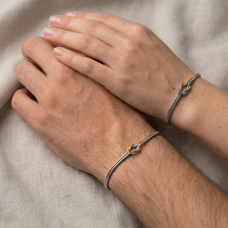 May include: Two silver bracelets, each featuring a knot design, are worn on wrists. The bracelets are simple and elegant, with a polished finish. The background is a neutral color, highlighting the jewelry.