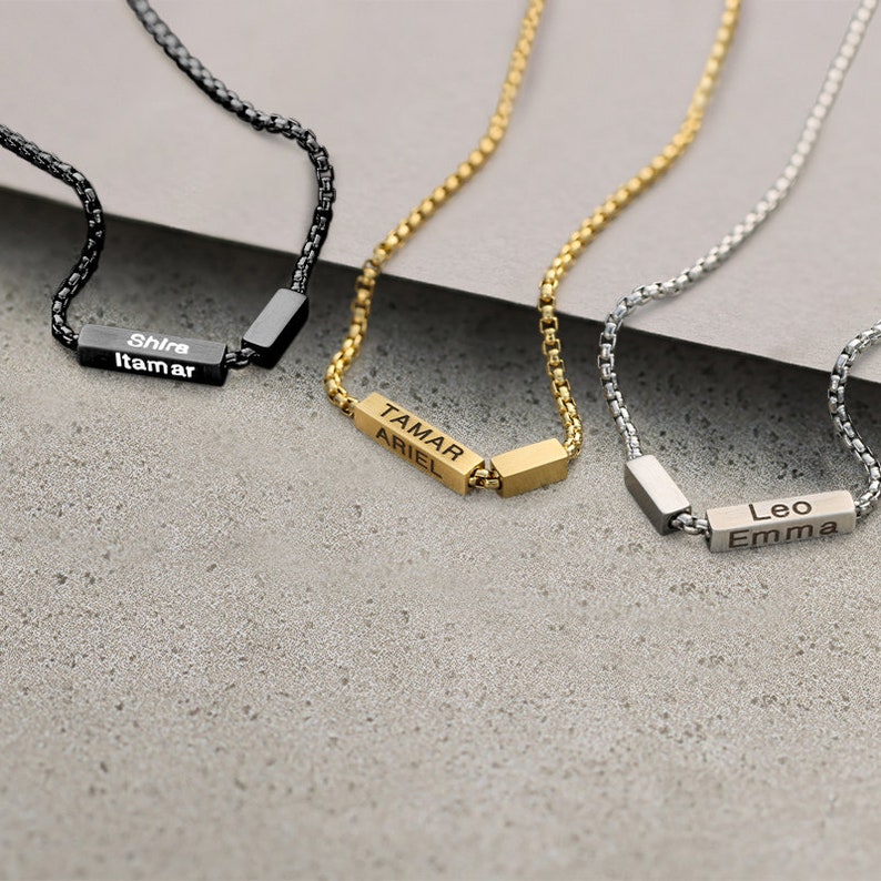 Puede incluir: Tres collares de cadena dorada, plateada y negra con etiquetas de nombre personalizadas. El collar dorado tiene los nombres "Tamar" y "Ariel" en la etiqueta. El collar plateado tiene los nombres "Leo" y "Emma" en la etiqueta. El collar negro tiene los nombres "Shira" y "Itamar" en la etiqueta.