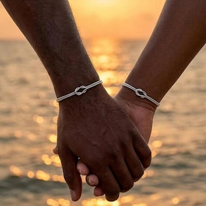 May include: Two silver knot bracelets are worn on wrists, with hands clasped together. The bracelets are simple, elegant, and feature a knot design. The background is a blurred view of the ocean at sunset, with warm orange and yellow tones.