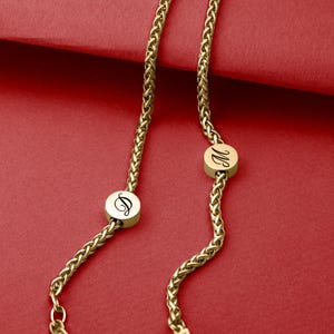 May include: Gold-colored necklace with a wheat chain design. The necklace features two circular pendants with the letters "D" and "M" in a cursive font. The background is a solid red color.