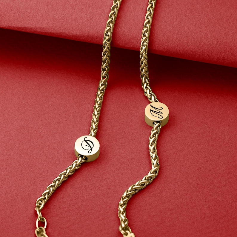 May include: Gold-colored necklace with a wheat chain design. The necklace features two circular pendants with the letters "D" and "M" in a cursive font. The background is a solid red color.