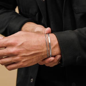 May include: Two silver bracelets are worn on a wrist. One is a thin, flat band, and the other is a delicate chain. The bracelets are displayed against a dark background, highlighting their metallic sheen. The jewelry is simple and modern.