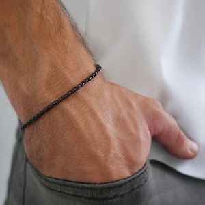 Puede incluir: Una pulsera trenzada negra se lleva en la muñeca. La pulsera es de metal oscuro y tiene un diseño tejido. La persona lleva una camisa blanca y pantalones grises. La pulsera es un accesorio elegante.