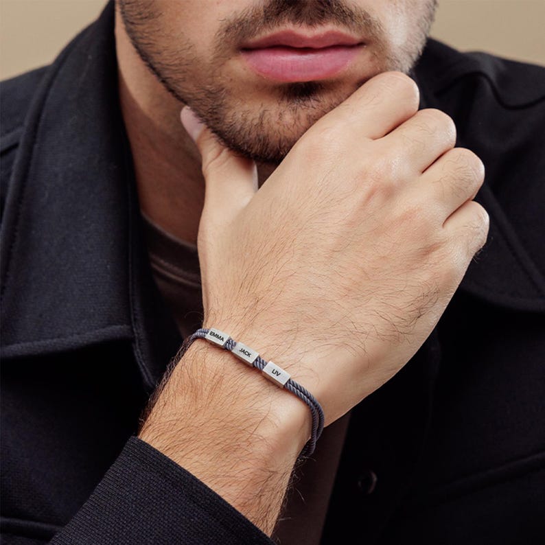 May include: A gray braided bracelet with silver beads that have the names "Emma", "Jack", and "Liv" engraved on them.