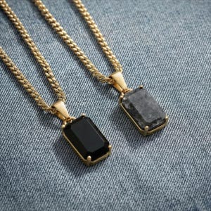 May include: Two gold-colored chain necklaces with rectangular pendants. One pendant is black, the other is gray. The pendants are set in gold-colored frames. The necklaces are displayed on a blue denim surface.
