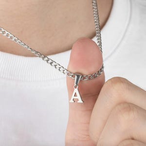 Puede incluir: Collar plateado con una cadena de eslabones y un colgante con forma de la letra "A". El colgante es pequeño y cuelga de un pequeño lazo sujeto a la cadena. El collar se muestra sobre un fondo blanco.