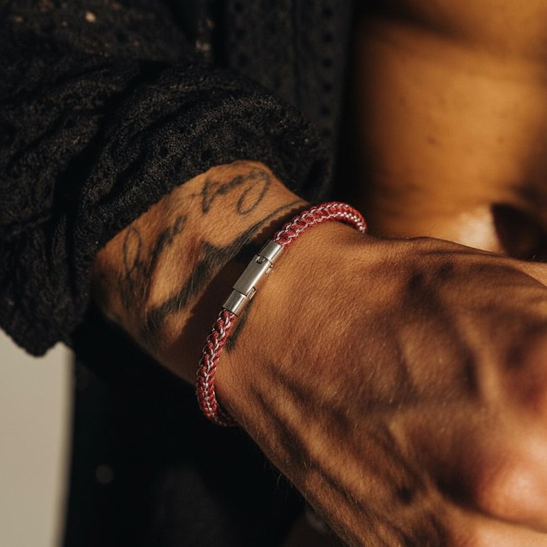 May include: A red and white braided bracelet with a silver clasp is worn on a wrist. The bracelet is made of a woven material. The wrist has a tattoo. The person is wearing a black shirt.