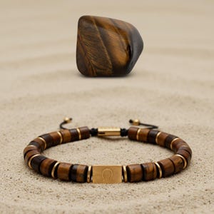 May include: A beaded bracelet with brown and gold accents and a gold-colored clasp featuring the Omega symbol. A polished, brown tiger's eye stone sits above the bracelet on a sandy surface. The bracelet is made of wooden beads.