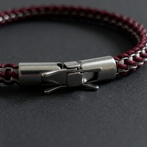 May include: Close-up of a bracelet with a burgundy and silver chain design. The bracelet features a stainless steel clasp. The chain is a woven pattern of burgundy and silver links. The clasp is a secure, hinged design.