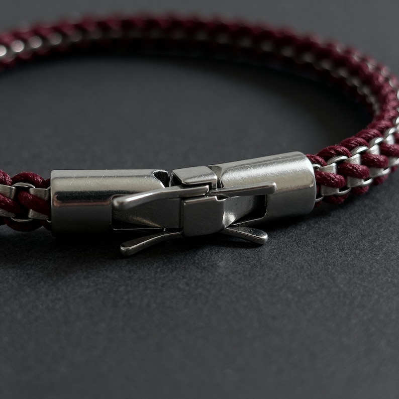May include: Close-up of a bracelet with a burgundy and silver chain design. The bracelet features a stainless steel clasp. The chain is a woven pattern of burgundy and silver links. The clasp is a secure, hinged design.