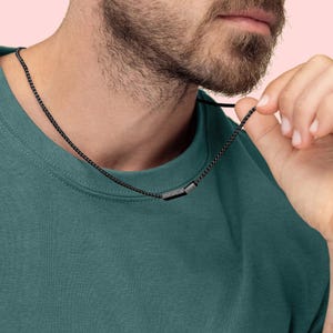 Puede incluir: Un collar de cadena negra con un pequeño colgante rectangular.