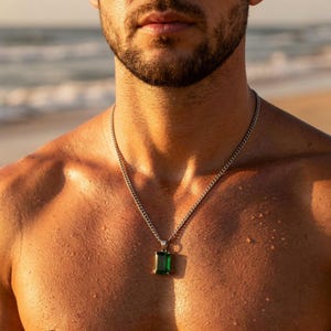May include: A silver chain necklace with a rectangular emerald green pendant. The pendant has a gold-colored border. The necklace is worn on a person's bare chest, with water droplets visible on the skin.