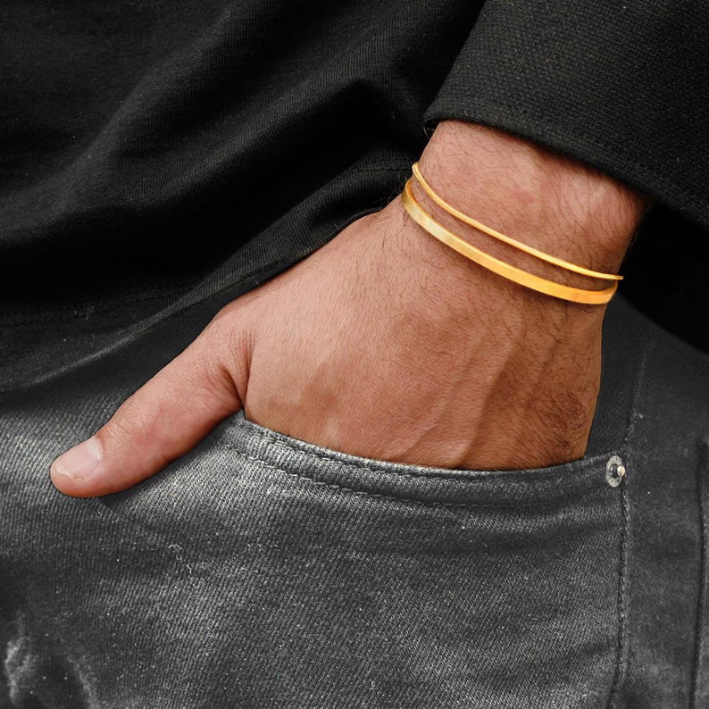 May include: Two gold-colored, thin, flat bracelets are worn on a wrist. The bracelets are stacked. The person is wearing a black long-sleeve shirt and gray jeans. The person's hand is in the pocket of the jeans.